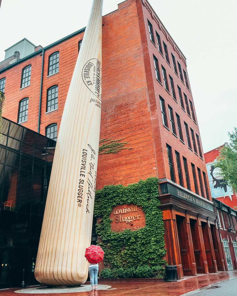 Girl with umbrella standing in the rain at the foot of a Giant Baseball Bat at Louisville Slugger Museum on Museum Row in Louisville Kentucky
