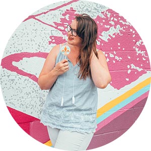 Lets Go Louisville creator Lia Garcia against a Butchertown mural eating a fleur de lis lollipop