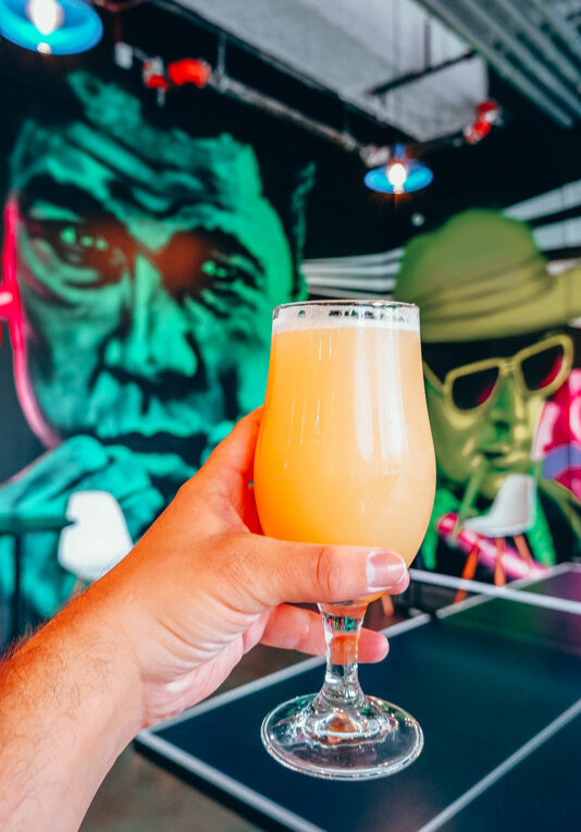 lo-pitch hazy ipa against a Muhammed Ali and Hunter S Thompson murals at hi-wire brewing louisville